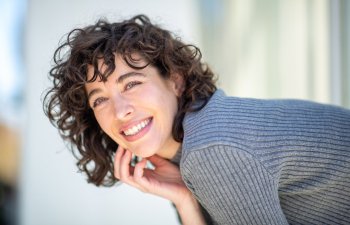 Person with curly hair and a gray sweater smiling and leaning slightly forward against a light background.